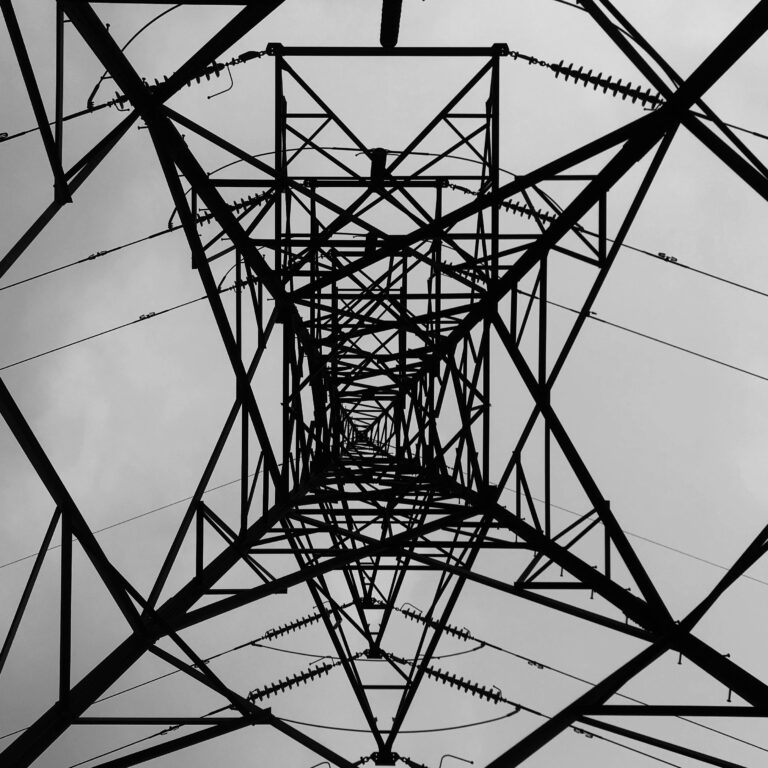 Black and white view from beneath a geometric steel power transmission tower.